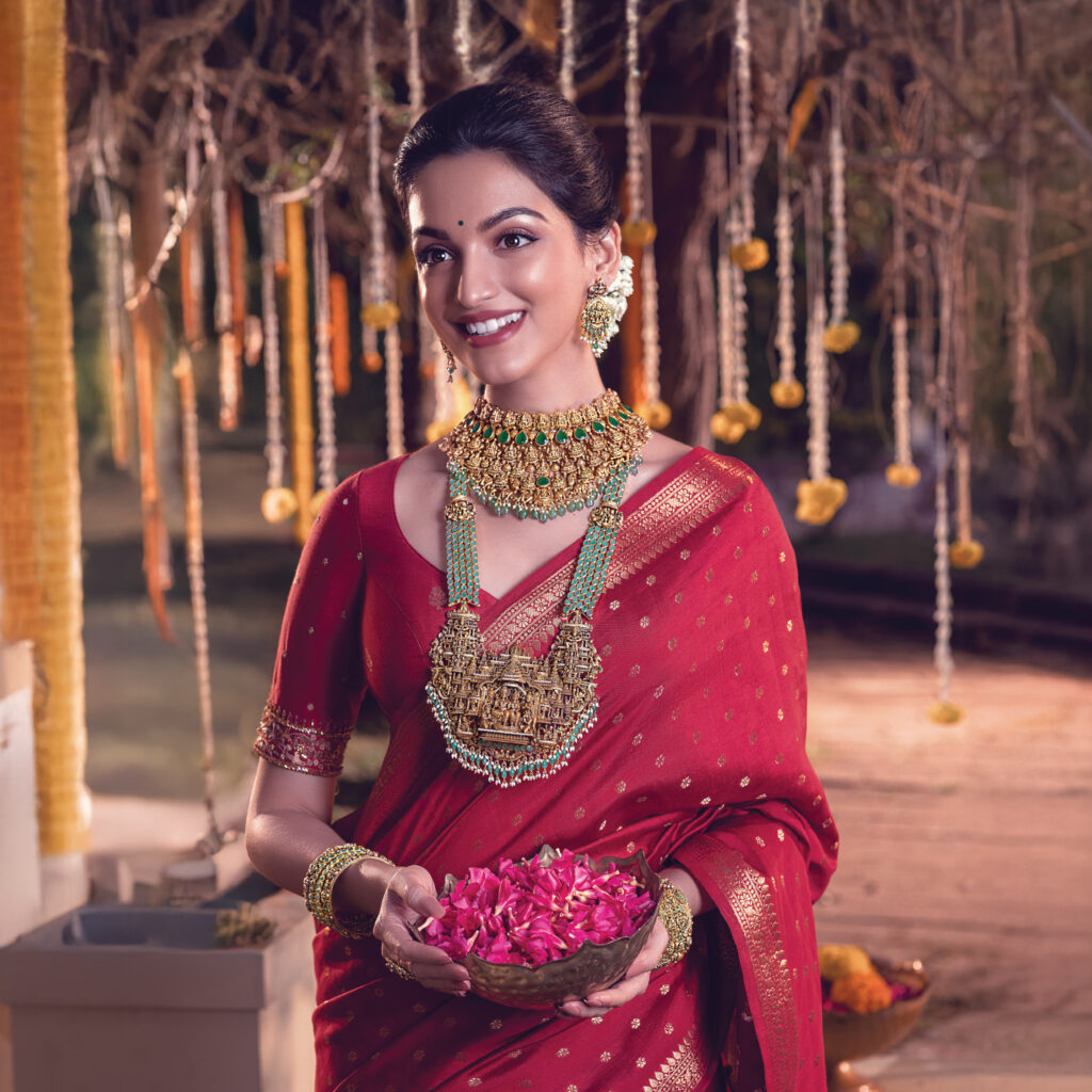 Bride wearing a handcrafted gold temple jewellery set with a Lakshmi motif long haram, emerald choker, earrings, and bangles from Regal Jewellers — a traditional South Indian bridal look in a red silk saree.